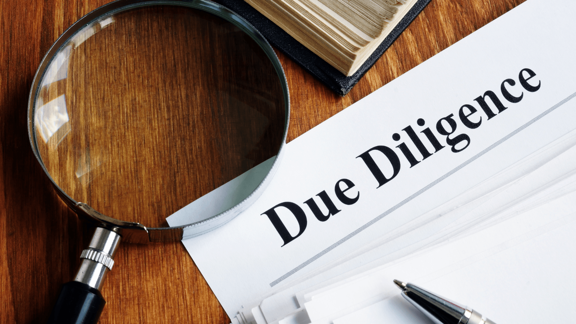 Magnifying glass over a document labeled 'Due Diligence' on a wooden desk, next to a pen and a stack of papers, symbolizing careful review and investigation