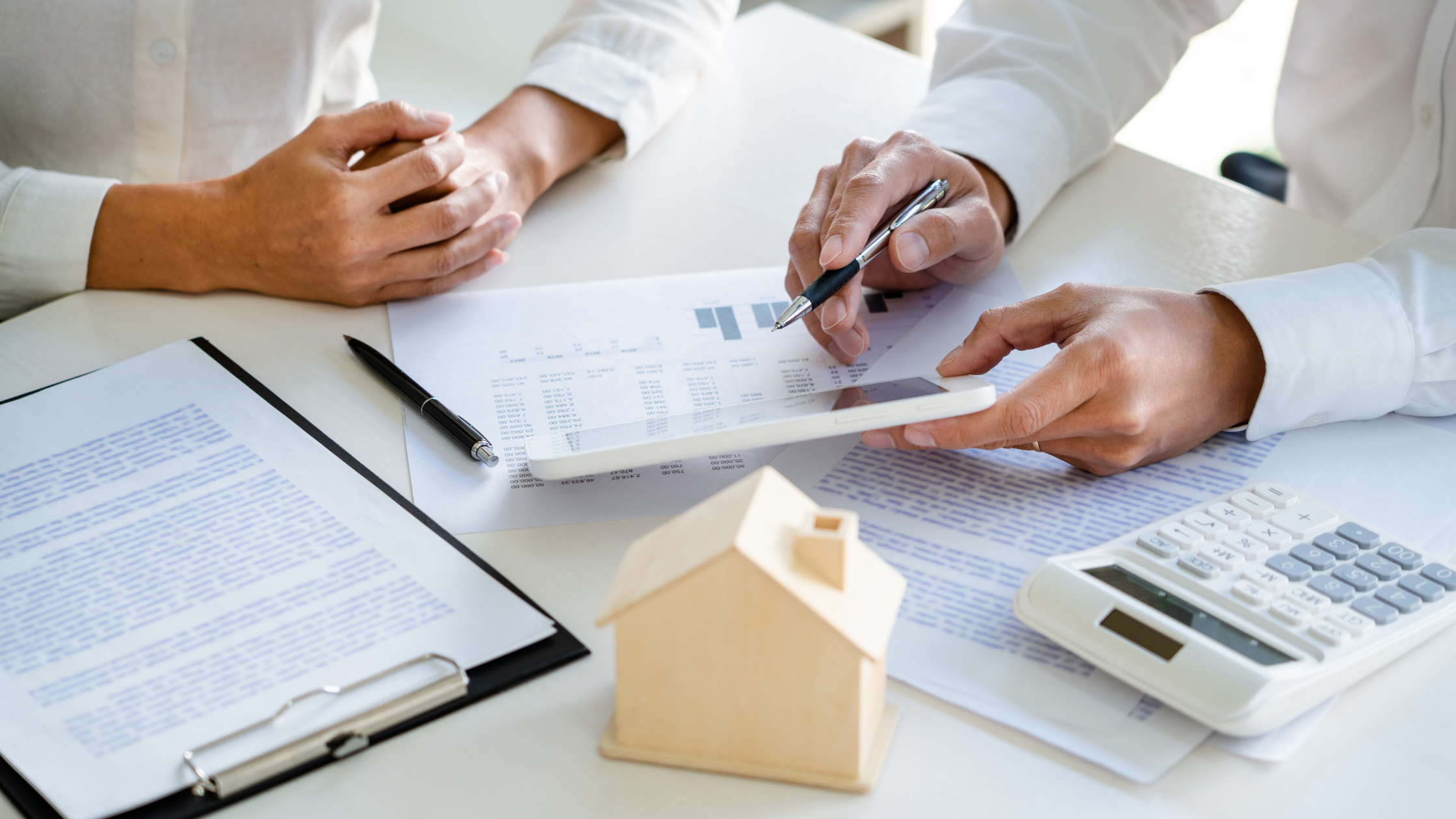 Business meeting discussing real estate documents and property financing, with a house model, calculator, contracts, and financial reports on a desk.