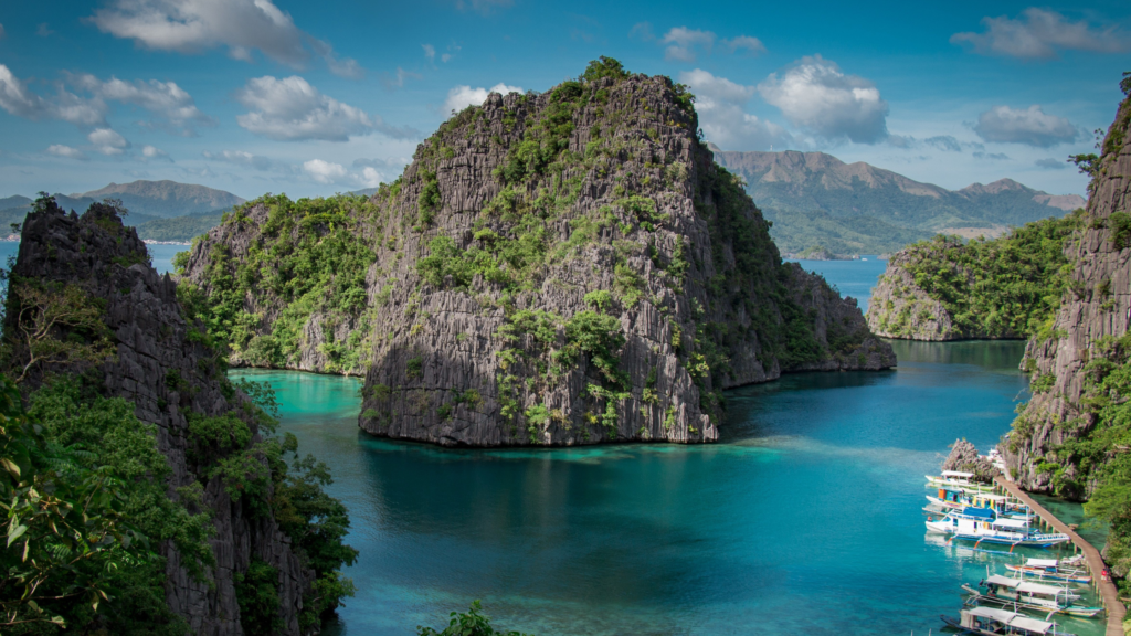 Scenic view of turquoise waters surrounded by dramatic limestone cliffs, lush greenery, and mountains in Palawan, Philippines, with boats docked along the shore.